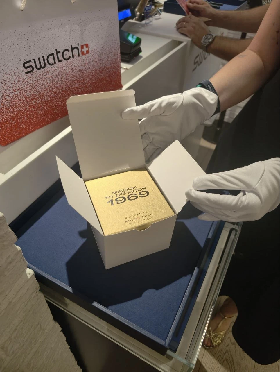 Eine Mitarbeiterin öffnet mit weissen Handschuhen die goldene Verpackung der MoonSwatch