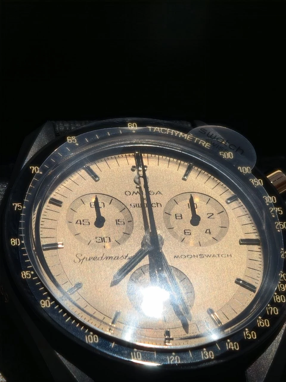 Nahaufnahme des goldfarbenen Zifferblatts und der schwarzen Tachymeter-Lünette der MoonSwatch